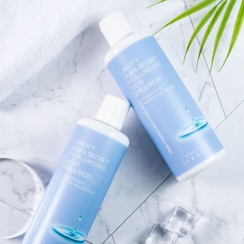 Two bottles of JIGOTT Hyaluronic Emulsion styled with marble surface and green props
