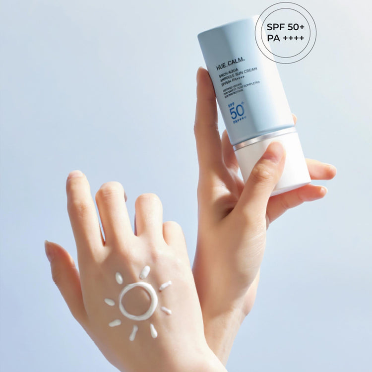Person holding Huecalm Birch Aqua Ampoule Sun Cream SPF 50+ PA++++ bottle; sun-shaped streak of lotion drawn on back of hand.